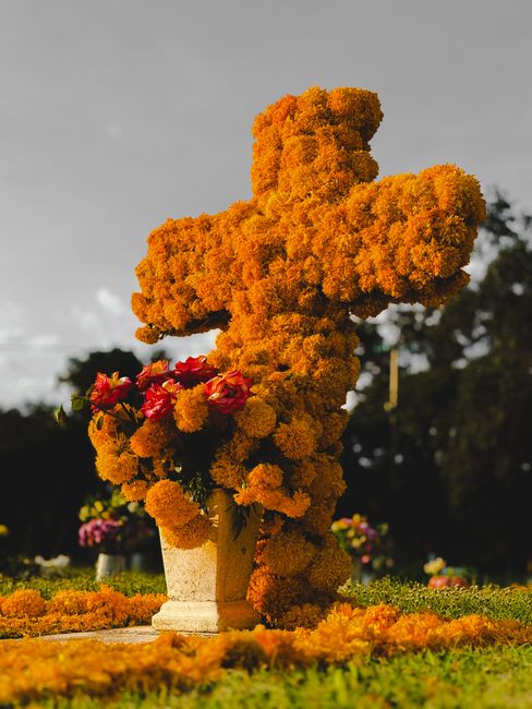 A floral cross memorial arrangement composed of densely packed orange marigold flowers, creating a textured and vibrant surface. The cross sits in a stone urn placed on a grassy area, with a layer of