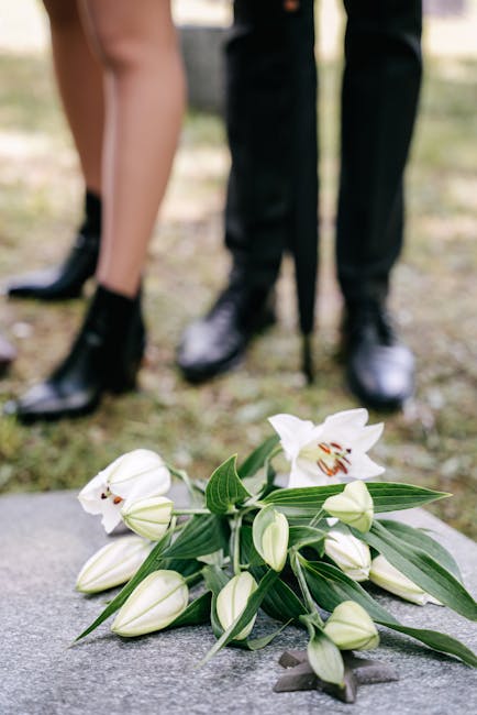 A bouquet of white lilies with large petals and prominent stamens, accompanied by closed white buds and long, slender green leaves, rests on a concrete surface. The flowers are fresh and vibrant, with