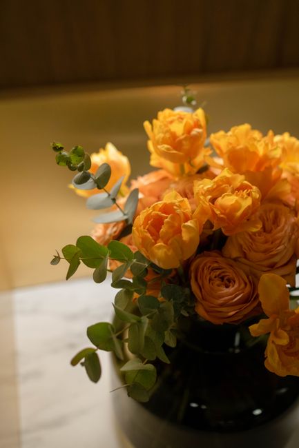 A floral arrangement in a dark, glossy vase featuring vibrant, double-blossomed orange roses and ranunculus with ruffled petals. The bouquet is accented with sprigs of eucalyptus leaves, providing a c