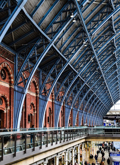 The image showcases the interior of Kings Cross Station, featuring an expansive arched metal and glass roof supported by intricate blue steel frameworks. Red brick walls with arched windows line the s