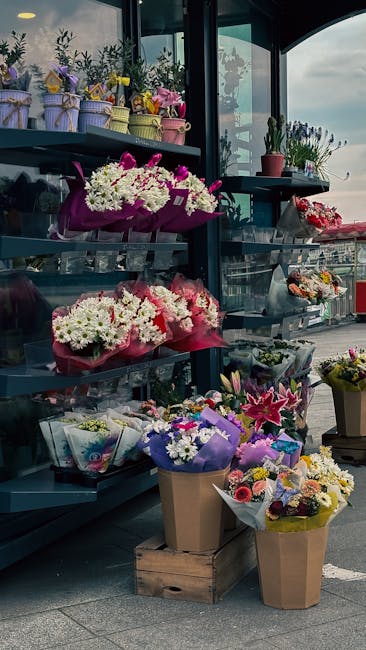 An outdoor display of floral arrangements with various bouquets of fresh flowers, including white daisies, pink lilies, and purple irises, arranged in colorful paper and plastic wrapping. Some bouquet