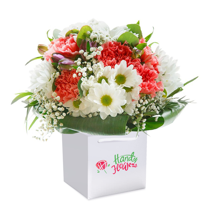 Presented against a bright, neutral backdrop, this exuberant arrangement embodies the dynamic spirit of Kings Cross. Expertly set within an immaculate white square gift bag, the bouquet overflows with lush coral, peach, and deep red carnations-each bloom layered with ruffled, almost feathery petals, creating a rich and tactile texture you can almost feel. Vibrant white daisy-like chrysanthemums, punctuated with striking yellow-green centers, add a note of freshness and energy, reminiscent of springtime strolls through Lewis Cubitt Park. Delicate clusters of white baby's breath weave seamlessly between the main florals, providing airy contrast and softening the scene. Abundant, glossy green foliage frames the flowers, with broad leafy arcs forming an elegant, natural collar around the base. A few partially closed buds, tinged with gentle purple hues, peek from the side, offering a sense of anticipation-like the promise of new blooms spotted in a Bloomsbury garden. The bouquet is arranged into a full, rounded formation, its effortless grace reminiscent of skilled floristry at its best. The white carrier bag, practical yet stylish with sturdy ribbon handles, makes the bouquet perfect for gifting on a walk through Pancras Square or for delivery to a friend along York Way. The photo's clarity and artful composition underscore the arrangement's freshness and sophistication, capturing the essence of a thoughtful locally crafted gift-ideal for celebrating special milestones, from anniversaries to heartfelt thank-yous within the vibrant community of Kings Cross.